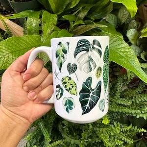 May include: White ceramic mug with a green and white leaf pattern. The mug has a handle and is filled with a white liquid.