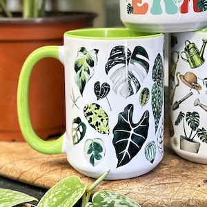 May include: White ceramic mug with a green handle and interior, adorned with various watercolor-style leaf illustrations. The mug is part of a set, with other mugs featuring botanical designs. The overall theme is plants and gardening.