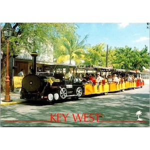 Könnte beinhalten: Eine Vintage-Postkarte mit einem schwarz-gelben Touristen-Zug, der durch eine Straße in Key West fährt. Der Zug ist voller Passagiere und hat einen dekorativen Schornstein. Der Text "KEY WEST" ist rot am unteren Bildrand aufgedruckt.