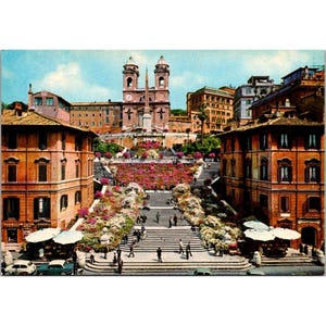 May include: Vintage postcard featuring the Spanish Steps in Rome, Italy. The image shows the wide staircase adorned with flowers, leading up to a church. Buildings with awnings and cars are visible at the base of the steps, with a blue sky overhead.
