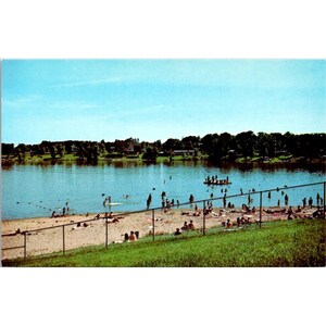May include: Vintage postcard featuring a lakeside beach scene. People are swimming and sunbathing on the sandy shore. A fence separates the beach from a grassy area. The sky is a clear blue, and trees line the far shore.