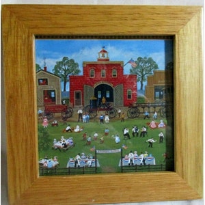 May include: A framed painting depicting a small town scene with a red brick fire station, a fire truck, and a group of people gathered for a picnic. The painting is in a rustic wooden frame.