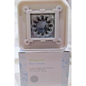 Puede incluir: Un punzón de papel de plástico blanco con una matriz de metal plateado en forma de flor. El punzón está en su caja, que es blanca con un patrón floral azul y verde. La caja dice "Blossom Place 'n' Punch" y tiene texto en francés, alemán y japonés.