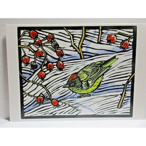 Op de afbeelding: Een linosnede van een vogel met groene en rode veren op een tak met rode bessen. De achtergrond toont een besneeuwd landschap met blauwe en witte tinten, omlijst door een zwarte rand.