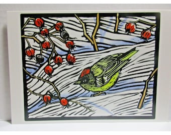 May include: A linocut print of a bird with green and red plumage perched on a branch with red berries. The background features a snowy landscape with blue and white tones, framed by a black border.