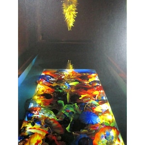 Puede incluir: Una instalación artística de vidrio colorida. Una estructura rectangular, similar a una mesa, está llena de piezas de vidrio superpuestas en rojo, naranja, amarillo, azul y verde. Una escultura de vidrio amarillo cuelga encima.