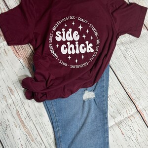 May include: A maroon t-shirt with a white circle graphic that says "side chick" with stars and a list of side dishes around the circle.
