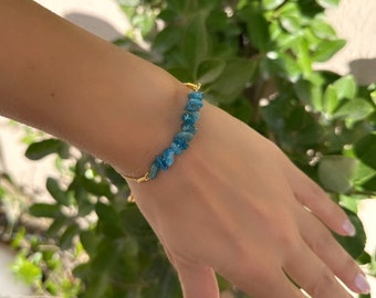 May include: A gold-toned chain bracelet featuring a series of small, irregular, bright blue apatite gemstone chips arranged in a row. The bracelet includes a delicate metallic chain and a secure clasp mechanism, showcasing a vibrant blue colour.