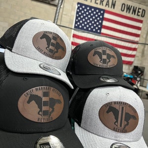 May include: Four black and grey baseball caps with a brown leather patch that says "United States Navy Chief Warrant Officer" and "Retired". The caps are arranged in a row with the American flag in the background. The flag says "Veteran Owned".