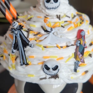 Puede incluir: Un cupcake tem&aacute;tico de Halloween con glaseado blanco, decorado con chispas y figuras de personajes de The Nightmare Before Christmas. La base es una taza con forma de calabaza con una cara de Jack Skellington.