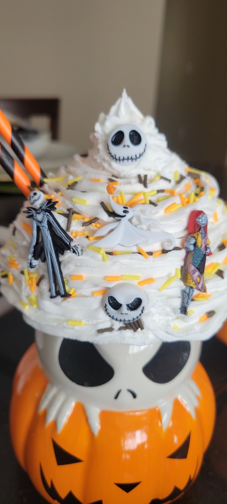 Puede incluir: Un cupcake tem&aacute;tico de Halloween con glaseado blanco, decorado con chispas y figuras de personajes de The Nightmare Before Christmas. La base es una taza con forma de calabaza con una cara de Jack Skellington.