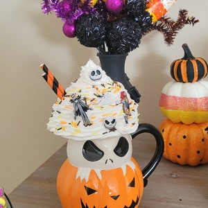 Puede incluir: Una taza de cer&aacute;mica con tem&aacute;tica de Halloween con forma de la cabeza de Jack Skellington, cubierta con crema batida y figuritas de personajes. La taza es naranja y blanca con un asa negra. En el fondo hay caramelos de ma&iacute;z decorativos y bolas de purpurina moradas.