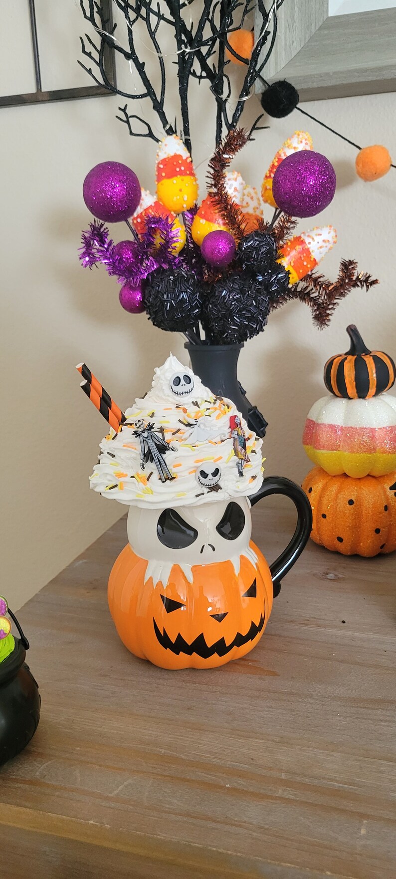 Puede incluir: Una taza de cer&aacute;mica con tem&aacute;tica de Halloween con forma de la cabeza de Jack Skellington, cubierta con crema batida y figuritas de personajes. La taza es naranja y blanca con un asa negra. En el fondo hay caramelos de ma&iacute;z decorativos y bolas de purpurina moradas.