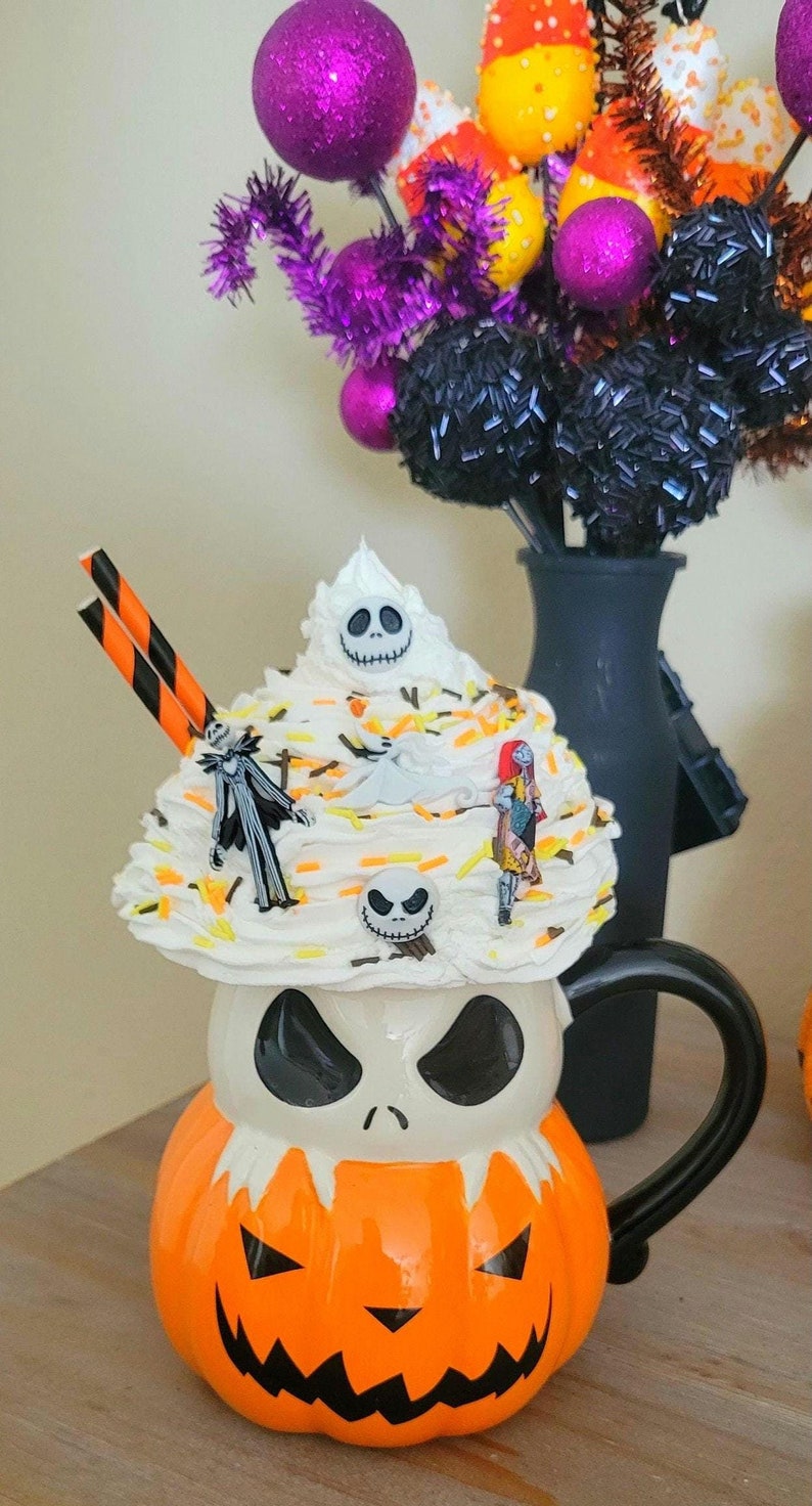Puede incluir: Una taza tem&aacute;tica de Halloween con forma de calabaza con una cara de Jack Skellington. La taza est&aacute; cubierta con crema batida, figuritas de personajes y chispas. En el fondo hay adornos decorativos de Halloween y un jarr&oacute;n negro.