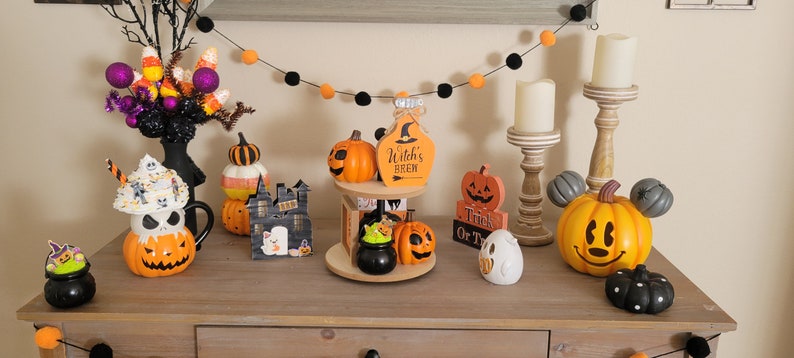 Puede incluir: Una colecci&oacute;n de decoraciones tem&aacute;ticas de Halloween, que incluyen figuras con forma de calabaza, una botella de poci&oacute;n de bruja y una calabaza de Mickey Mouse. La exhibici&oacute;n tambi&eacute;n presenta una guirnalda negra con pompones naranjas y negros y un &aacute;rbol decorativo con caramelos.