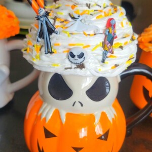 Puede incluir: Una taza de Halloween con forma de calabaza con la cara de Jack Skellington. La taza est&aacute; cubierta con crema batida y figuras de personajes de Pesadilla antes de Navidad, con pajitas naranjas y negras.