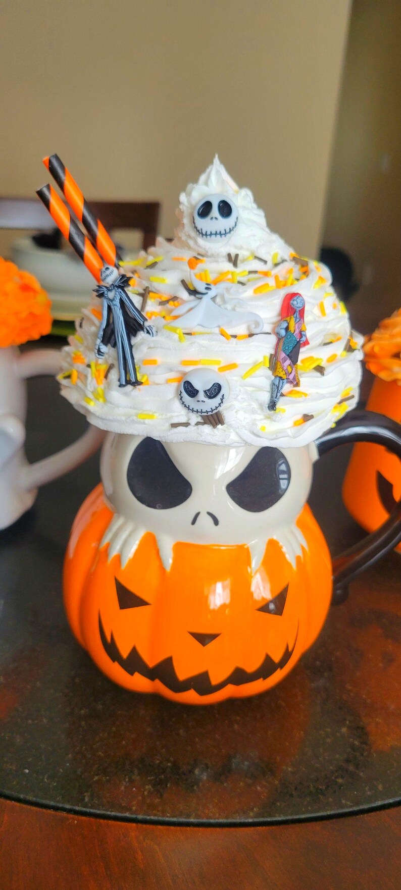 Puede incluir: Una taza de Halloween con forma de calabaza con la cara de Jack Skellington. La taza est&aacute; cubierta con crema batida y figuras de personajes de Pesadilla antes de Navidad, con pajitas naranjas y negras.