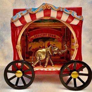 May include: A red and white circus wagon with a gold elephant figurine inside. The wagon has a sign that says "Greatest Show" and black wheels with gold accents.