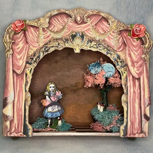 May include: A decorative wooden diorama featuring a theatrical stage frame with pink curtains and red roses. Inside, a cutout of a girl in a blue dress holds a piglet, while a blue cat rests in a pink tree above green moss and small mushrooms.