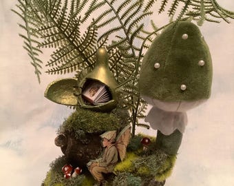 May include: A miniature fairy figurine sitting on a moss-covered log, reading a book inside a golden birdhouse. A green velvet mushroom with white pearl accents sits beside the log. A green fern branch hangs above the scene.