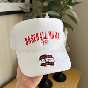 May include: White baseball cap with a red embroidered logo that says "BASEBALL MAMA" with a red bow. The cap has a black mesh back and a white label that says "OTTO WORLD FAMOUS FOAM FRONT OSFM TRUCKER DURABLE SNAP CLOSURE".
