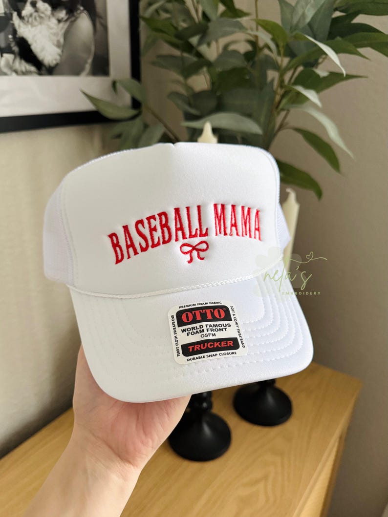May include: White baseball cap with a red embroidered logo that says "BASEBALL MAMA" with a red bow. The cap has a black mesh back and a white label that says "OTTO WORLD FAMOUS FOAM FRONT OSFM TRUCKER DURABLE SNAP CLOSURE".