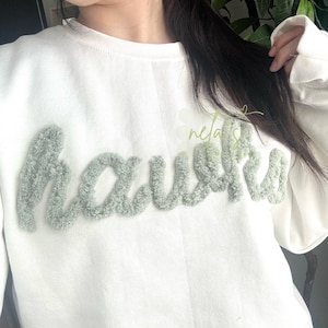 May include: White sweatshirt with the word "haunts" in a fuzzy, light green script across the chest. The sweatshirt has a crew neck and long sleeves. The word "nela's" is written in a light green script in the upper right corner.