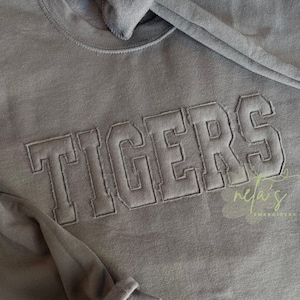 May include: A gray sweatshirt with the word "TIGERS" embroidered across the chest in a block letter style. The sweatshirt has a crew neck and a soft, textured appearance. The embroidery is in a matching gray color.