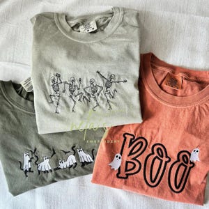 May include: Three t-shirts in shades of green and orange, each featuring Halloween-themed embroidery. One shirt has dancing skeletons, another has cats and ghosts, and the third says "Boo" with ghost accents. The shirts are folded and displayed on a white surface.