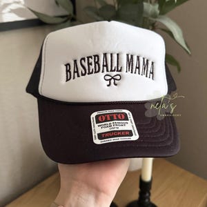 May include: A black and white trucker hat with a black mesh back. The front of the hat is white and has the text "BASEBALL MAMA" embroidered in black with a black bow. The hat has a black label that says "OTTO WORLD FAMOUS FOAM FRONT OSFM TRUCKER DURABLE SNAP CLOSURE".