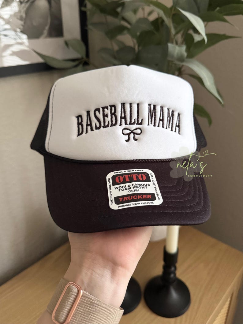 May include: A black and white trucker hat with a black mesh back. The front of the hat is white and has the text "BASEBALL MAMA" embroidered in black with a black bow. The hat has a black label that says "OTTO WORLD FAMOUS FOAM FRONT OSFM TRUCKER DURABLE SNAP CLOSURE".