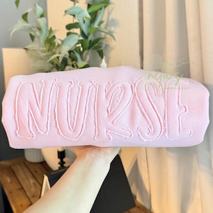 May include: A light pink rectangular pillow with the word "NURSE" embroidered in a matching light pink thread. The pillow is held up in front of a blurred background with greenery.