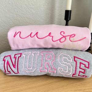 May include: A light pink sweatshirt with the word nurse embroidered in a dark pink cursive script, stacked on a heather gray sweatshirt featuring the word NURSE in large, block-style letters with a dark pink outline and light pink fill.
