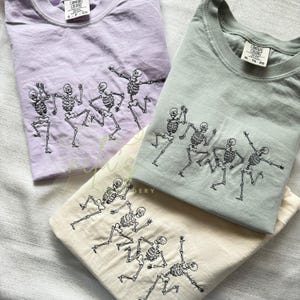 May include: Three folded t-shirts in lavender, sage green, and cream. Each shirt features a black embroidered design of dancing skeletons. The shirts are made of soft material and are suitable for casual wear.
