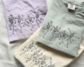 May include: Three folded t-shirts in lavender, sage green, and cream. Each shirt has a black embroidered design of dancing skeletons. The shirts are made of soft material and are suitable for casual wear.