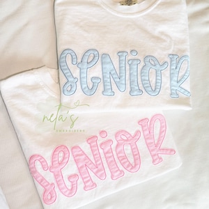 May include: Two white t-shirts featuring the word SENIOR in large, embroidered, bubble-style lettering. One shirt displays the text in light blue, while the other shirt features the text in light pink. Both shirts have a crew neckline.