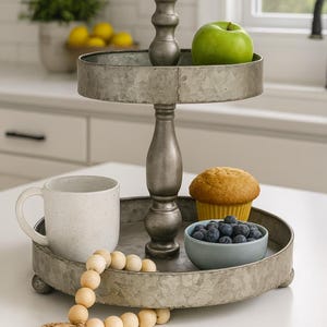 May include: A two-tiered metal serving tray with a handle, holding a green apple, a muffin, blueberries, and a mug. A wooden bead garland is draped on the bottom tier. The tray is on a white surface.