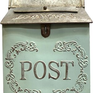May include: A light blue metal wall-mounted mailbox featuring a hinged lid with a metallic finish and embossed floral scrollwork. The front panel displays the word POST in large capital letters, framed by decorative embossed scroll patterns.