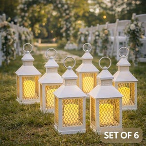 May include: Set of six white metal lanterns with a warm yellow glow, perfect for outdoor events. Each lantern features a diamond-patterned mesh and a handle. The text "SET OF 6" is visible in the bottom right corner.