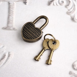 May include: A heart-shaped antique brass padlock with two matching keys. The lock has a striped design and a curved handle. The keys are attached to a ring. The objects are on a white surface with decorative embossed details.