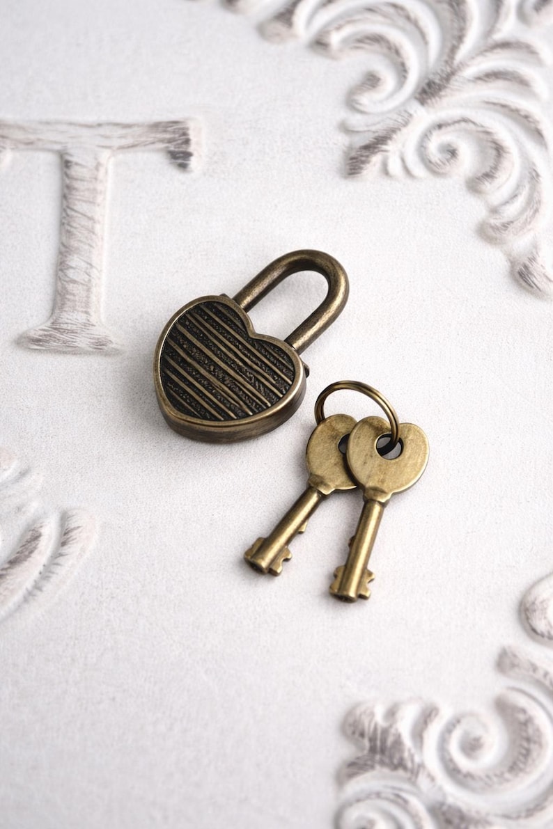 May include: A heart-shaped antique brass padlock with two matching keys. The lock has a striped design and a curved handle. The keys are attached to a ring. The objects are on a white surface with decorative embossed details.