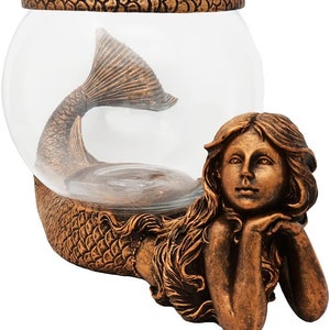 May include: A bronze-colored mermaid-themed fishbowl. The bowl is clear glass, with a mermaid tail inside. The mermaid figure is resting on her elbows, with long flowing hair. The base and rim of the bowl have a textured, scale-like design.