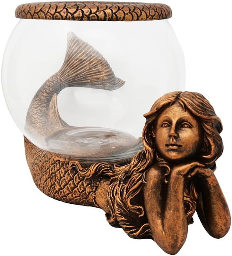 May include: A bronze-colored mermaid-themed fishbowl. The bowl is clear glass, with a mermaid tail inside. The mermaid figure is resting on her elbows, with long flowing hair. The base and rim of the bowl have a textured, scale-like design.