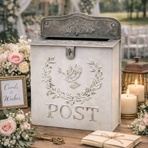 May include: A white metal post box with a decorative dove and wreath design. The word "POST" is embossed on the front. A key, cards, and candles are arranged on a wooden table, suggesting a wedding or event setting.