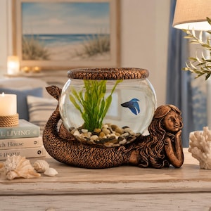 May include: A decorative fishbowl with a bronze mermaid base. The glass bowl contains a blue fish, green plants, and small pebbles. The mermaid is lying down, supporting the bowl with her arm. The bowl is topped with a textured bronze lid.