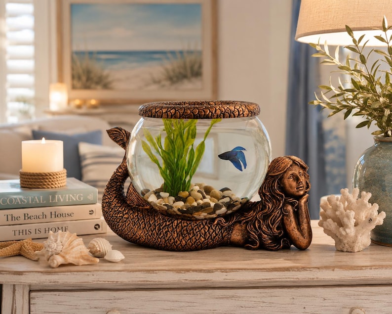 May include: A decorative fishbowl with a bronze mermaid base. The glass bowl contains a blue fish, green plants, and small pebbles. The mermaid is lying down, supporting the bowl with her arm. The bowl is topped with a textured bronze lid.