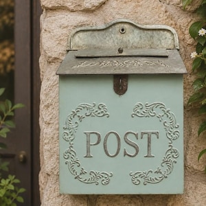 May include: A light mint green metal mailbox with a weathered finish. The front features the word POST in raised silver lettering, surrounded by an ornate embossed scrollwork border. It has a hinged lid and a decorative arched top panel.