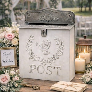 May include: A vintage-style wedding card box with a dove and wreath design, the word "POST" is embossed on the front. The box is made of a light-colored metal and sits on a wooden table with flowers and candles. The text "Wedding Card Box" is at the bottom.