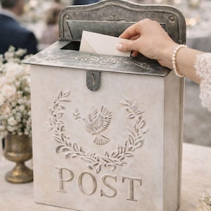 May include: A vintage-style mailbox with a dove and laurel wreath design. The word "POST" is embossed on the front. The mailbox is light beige with a metal lid and a hand is placing a card inside.