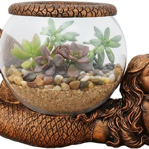May include: A bronze-colored mermaid figurine cradles a clear glass terrarium filled with succulents, sand, and small pebbles. The mermaid has long flowing hair and is resting her head on her hands. The terrarium is round with a decorative rim.